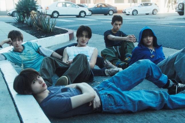 “Comeback concept photo of CORTIZ’s five members sitting and lying casually on a city sidewalk, wearing relaxed streetwear against an urban backdrop with parked cars and buildings, highlighting the group’s youthful and laid‑back vibe.