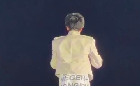 G‑Dragon from behind wearing the controversial light‑colored jacket and pants with the printed text, captured in a dark stage‑like setting.