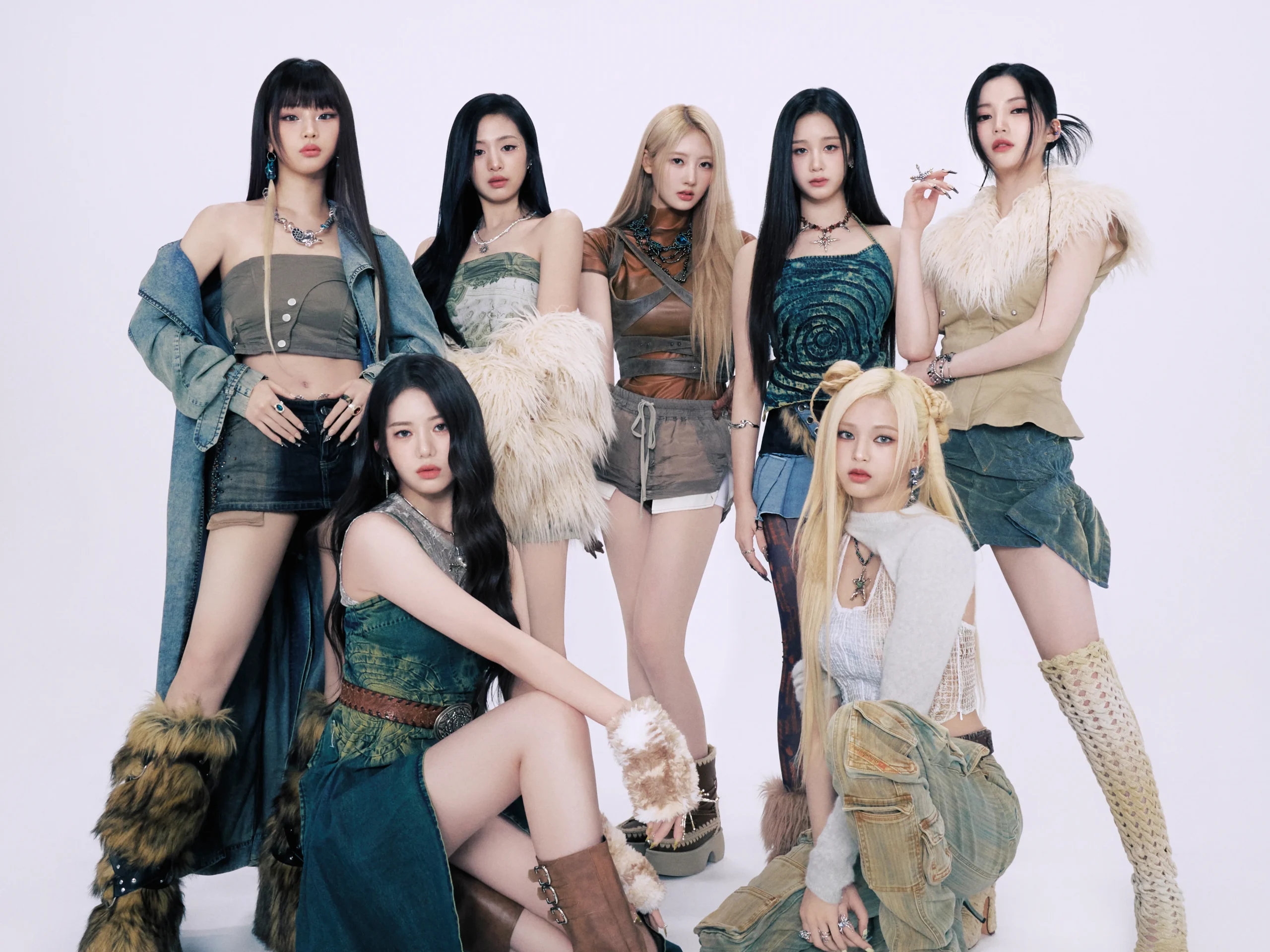 Promotional group photo of BABYMONSTER’s seven members standing against a light background, wearing modern fantasy‑inspired outfits with layered fabrics, faux fur, denim, and accessories, showcasing the group’s bold and stylish concept.