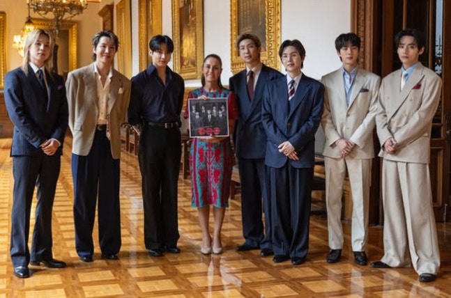 Group photo of BTS members Jimin, J-Hope, Jungkook, RM, Suga, Jin, and V standing with Mexico’s President Claudia Sheinbaum during an official meeting, captured in an ornate ceremonial room.