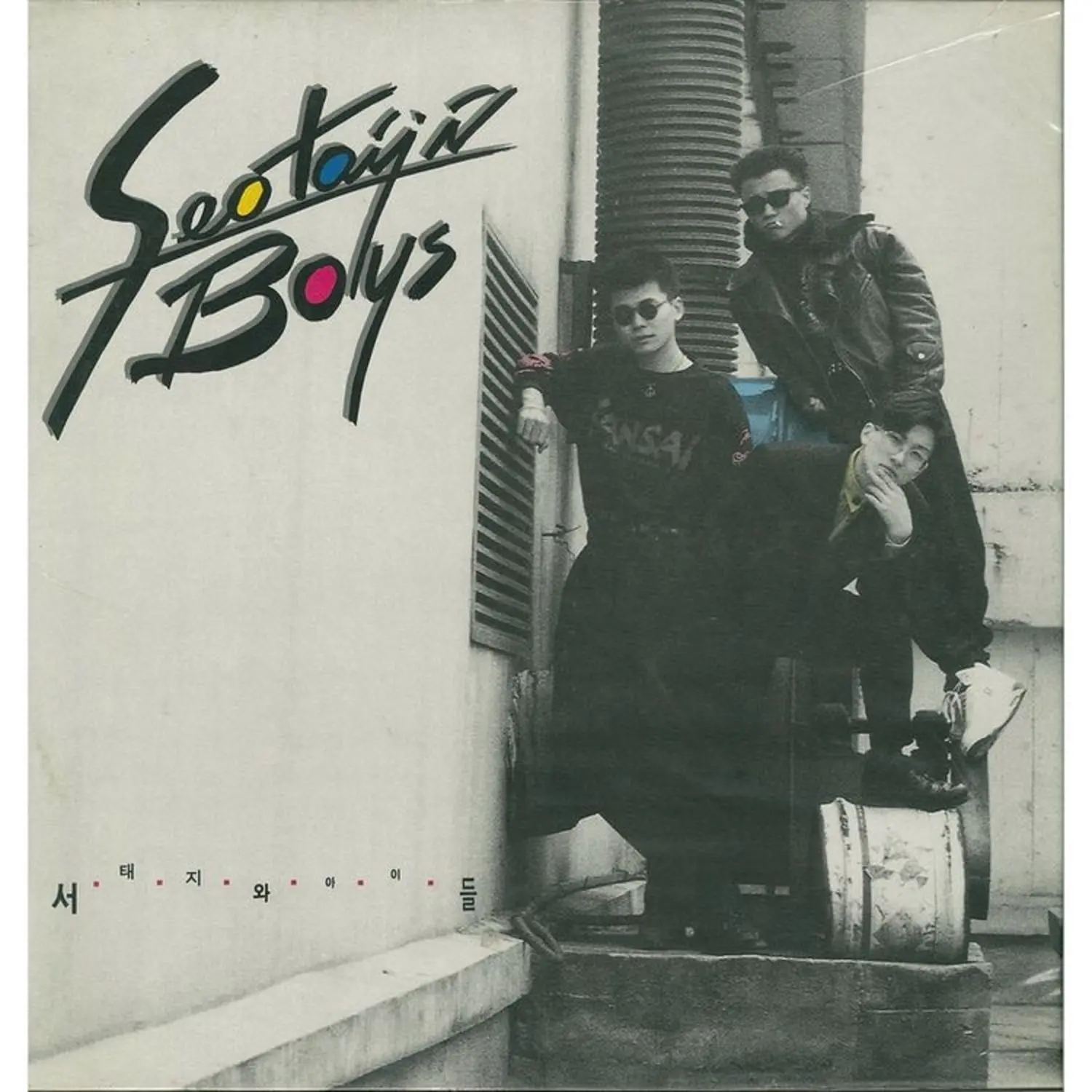 1992 debut album cover of Seo Taiji and Boys, featuring the three members posing in dark streetwear on an urban rooftop with industrial structures behind them; a landmark image in early K‑pop history with the stylized title ‘Seo Taiji & Boys’ and the Korean name ‘서태지와 아이들’ displayed prominently.