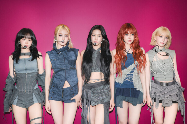 Promotional portrait of LE SSERAFIM’s five members standing against a bright magenta background, wearing futuristic performance outfits and headsets, showcasing the group’s bold and modern artist concept.