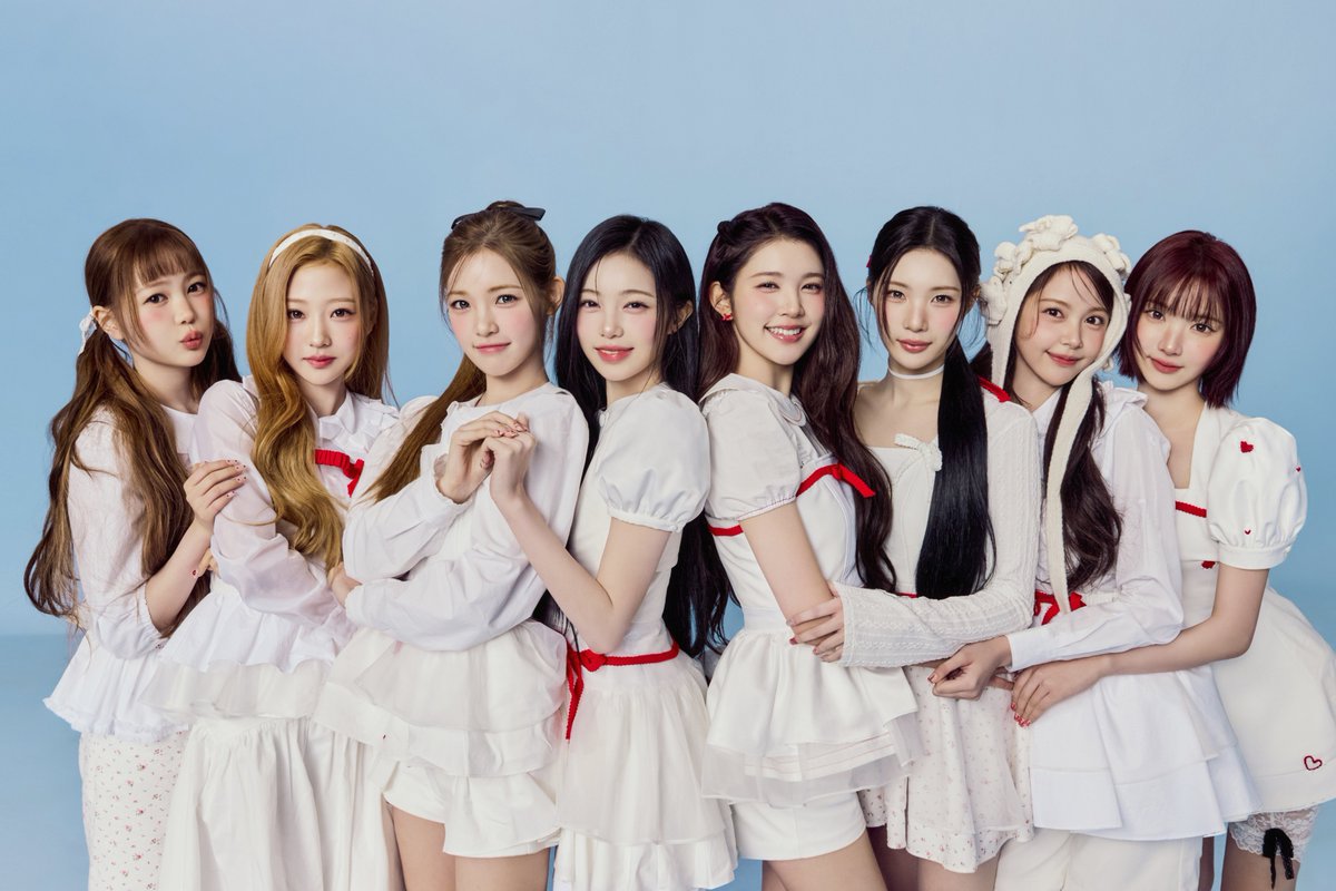 Group photo of Hearts2Hearts’ eight members standing together against a light blue background, wearing coordinated white outfits with red accents, highlighting the group’s bright and youthful concept