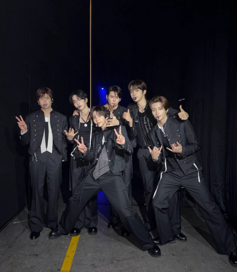 Backstage group photo of RIIZE’s six members wearing coordinated black performance outfits and headset microphones, posing together in a dimly lit stage area with a confident pre‑show energy.