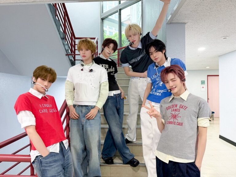 Group photo of TWS’s six members posing on an indoor staircase, wearing casual layered outfits and headset microphones, showcasing the group’s relaxed backstage performance concept.
