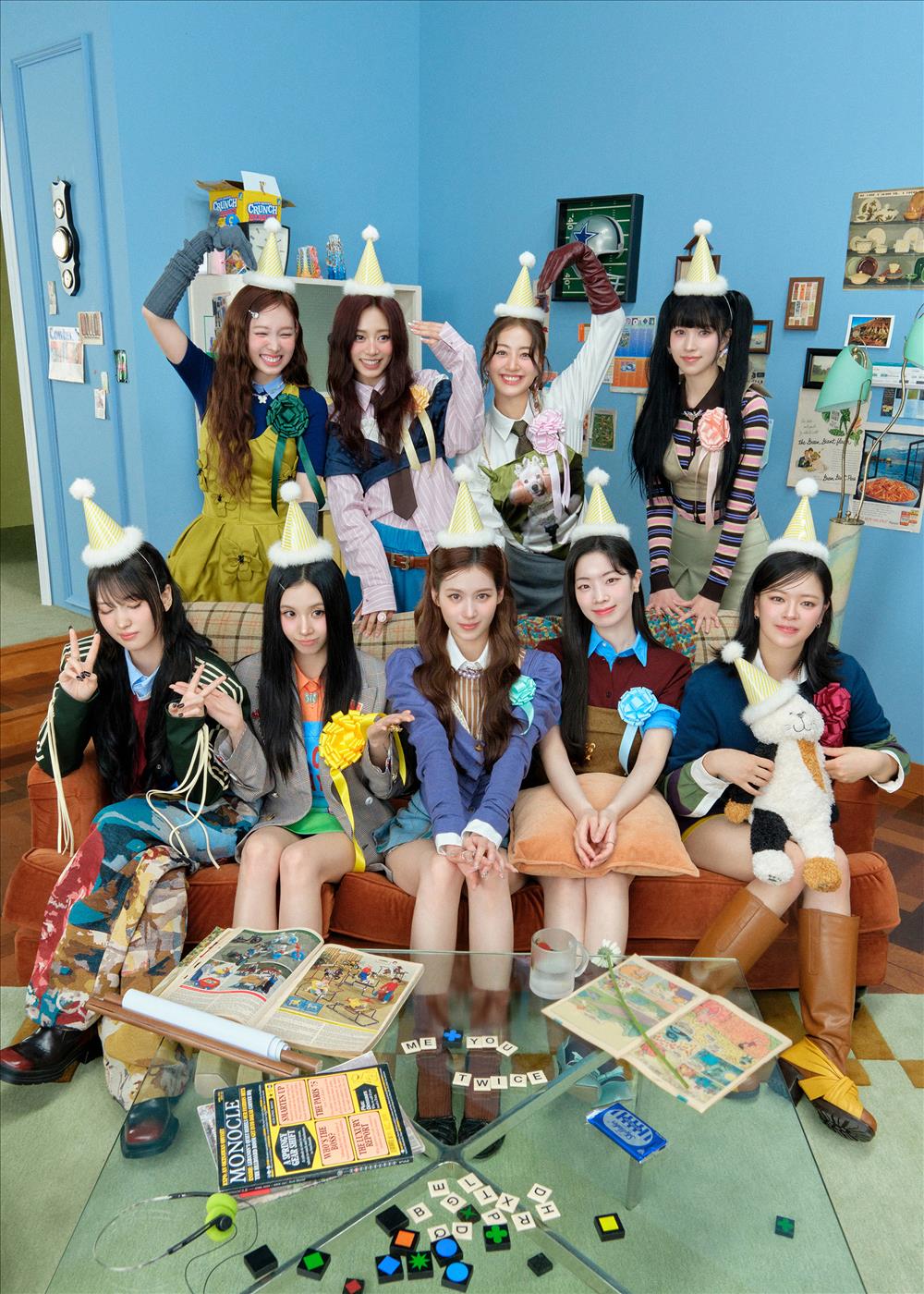 Group photo of TWICE’s nine members in a bright, playful room wearing party hats and colorful outfits, gathered around a table with magazines, Scrabble tiles spelling ‘ME YOU TWICE,’ and various props, creating a cheerful and celebratory concept.