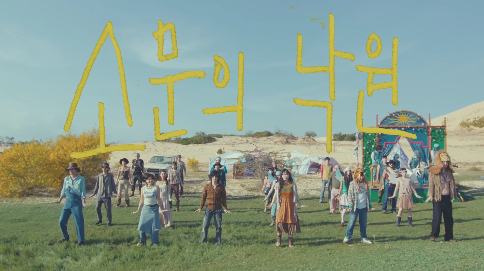 Promotional image for AKMU’s ‘Somunui Nakwons,’ featuring a colorful group gathered on a grassy field in front of desert‑like sand dunes, wearing eclectic costumes and standing near tents and a sun‑shaped structure, creating a surreal festival‑style concept.